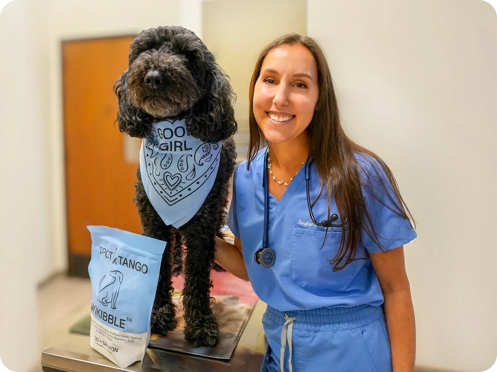 Spot & Tango Veterinary Advisor smiling beside a black curly-haired dog on an exam table next to a bag of UnKibble
